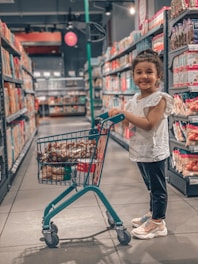 A young child stands in a grocery store aisle holding onto a small shopping cart filled with assorted groceries. The store shelves are stocked with various products, and the atmosphere is bright and organized. The child is smiling and wearing casual clothing.
