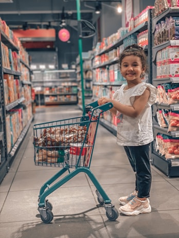 A young child stands in a grocery store aisle holding onto a small shopping cart filled with assorted groceries. The store shelves are stocked with various products, and the atmosphere is bright and organized. The child is smiling and wearing casual clothing.