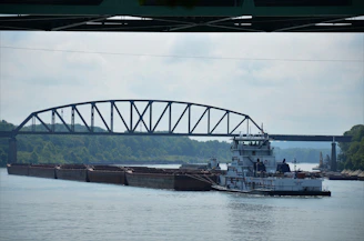 A river scene features a large barge carrying cargo, guided by a towboat beneath a steel truss bridge. The surrounding environment includes lush green trees lining the riverbank under a cloudy sky.