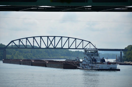 A river scene features a large barge carrying cargo, guided by a towboat beneath a steel truss bridge. The surrounding environment includes lush green trees lining the riverbank under a cloudy sky.