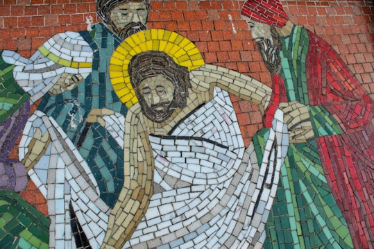 A mosaic depicting several figures, one with a halo, possibly representing a biblical scene. The figures are intricately constructed with small tiles in various colors, including reds, greens, yellows, and blues. The central figure is draped in white cloth and surrounded by others who appear attentive and somber.