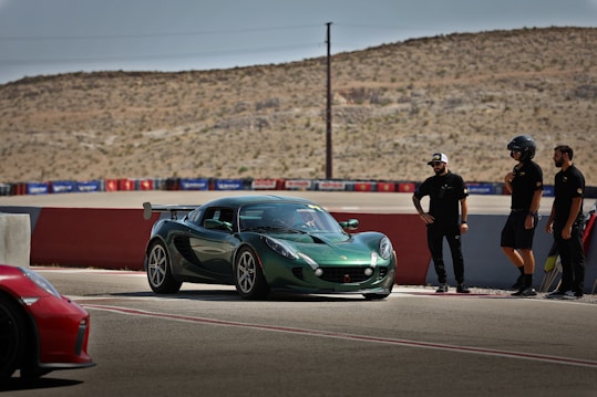 A green sports car is situated on a racetrack with a desert landscape in the background. Four individuals, some wearing helmets, stand nearby observing the vehicle. The track is bordered by a red and white barrier and there are advertisements along the fence.