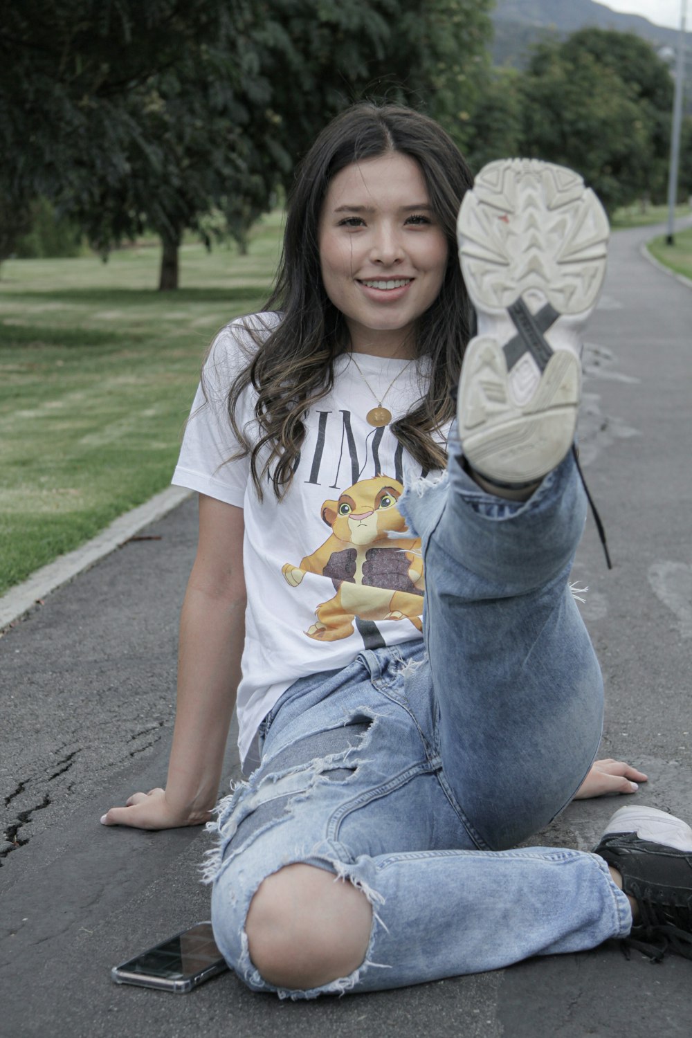 smiling girl in white t-shirt and blue denim jeans sitting on gray concrete pavement during