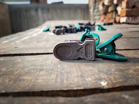 A close-up of a metal and plastic clip lying on a wooden surface. The clip is primarily green and silver, with a textured metal tip. In the background, there are blurred objects and a brick wall.