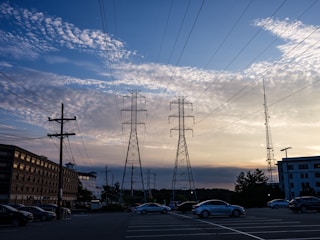 Power lines stretch across the sky with tall electricity pylons in the center. The sky displays a mix of blue and orange hues as the sun sets, with scattered clouds adding texture. In the foreground, a parking lot is visible with several parked cars, and buildings line the left side of the image.