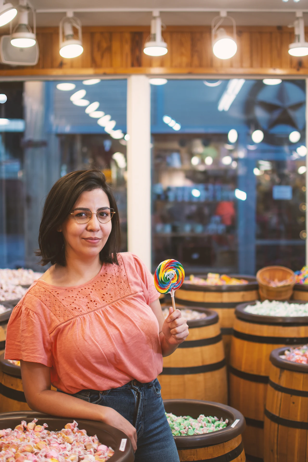 Woman in a coral top holds a rainbow lollipop among barrels of sweets in a brightly lit sweet shop.