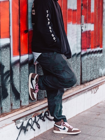 A person leans casually against a wall covered with graffiti. The individual is wearing a black hoodie with text on the sleeves, dark pants, and stylish sneakers featuring a mix of white, brown, and black colors.