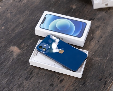 A new blue smartphone is partially removed from its white packaging, placed on a textured wooden surface. The phone's sleek, glossy finish and distinctive logo are visible, with a screen protector or insert showing a digital design. The opened box reveals additional packaging materials underneath.
