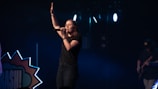 A female singer stands on a dimly lit stage, holding a microphone and raising one hand, conveying a sense of performance or worship. She is dressed in casual clothing, and the background includes stage lights and a partial view of a guitarist.