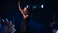 A female singer stands on a dimly lit stage, holding a microphone and raising one hand, conveying a sense of performance or worship. She is dressed in casual clothing, and the background includes stage lights and a partial view of a guitarist.