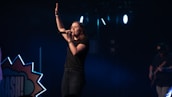 A female singer stands on a dimly lit stage, holding a microphone and raising one hand, conveying a sense of performance or worship. She is dressed in casual clothing, and the background includes stage lights and a partial view of a guitarist.