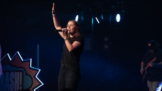 A female singer stands on a dimly lit stage, holding a microphone and raising one hand, conveying a sense of performance or worship. She is dressed in casual clothing, and the background includes stage lights and a partial view of a guitarist.