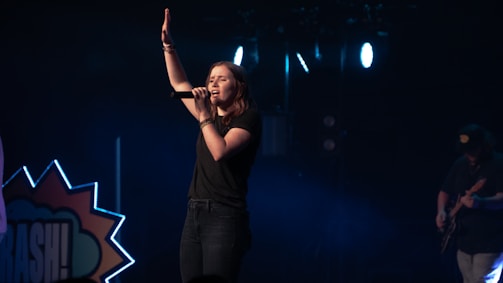 A female singer stands on a dimly lit stage, holding a microphone and raising one hand, conveying a sense of performance or worship. She is dressed in casual clothing, and the background includes stage lights and a partial view of a guitarist.