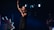 A female singer stands on a dimly lit stage, holding a microphone and raising one hand, conveying a sense of performance or worship. She is dressed in casual clothing, and the background includes stage lights and a partial view of a guitarist.