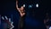 A female singer stands on a dimly lit stage, holding a microphone and raising one hand, conveying a sense of performance or worship. She is dressed in casual clothing, and the background includes stage lights and a partial view of a guitarist.