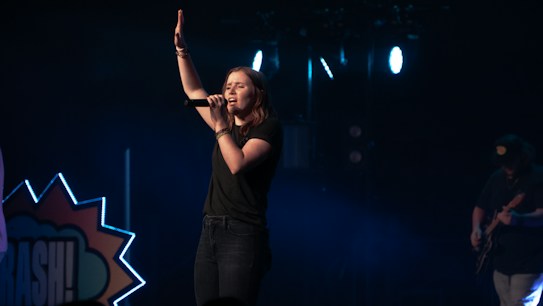 A female singer stands on a dimly lit stage, holding a microphone and raising one hand, conveying a sense of performance or worship. She is dressed in casual clothing, and the background includes stage lights and a partial view of a guitarist.