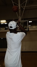 A person in a white shirt and cap is shooting a basketball towards a hoop in a dimly lit indoor gym. The basketball is in mid-air, just above the player's hands. The surroundings include a basketball court and visible hoop structure.