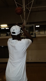 A person in a white shirt and cap is shooting a basketball towards a hoop in a dimly lit indoor gym. The basketball is in mid-air, just above the player's hands. The surroundings include a basketball court and visible hoop structure.