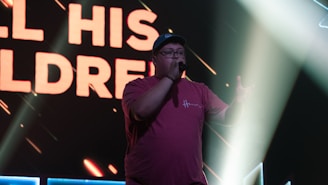 Person wearing a red t-shirt and cap, holding a microphone on a stage with bright lights and a large screen displaying bold text in the background.