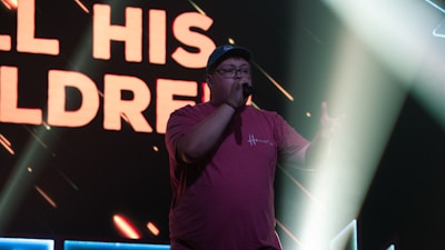 Person wearing a red t-shirt and cap, holding a microphone on a stage with bright lights and a large screen displaying bold text in the background.