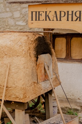 Outdoor scene showing a rustic, wood-fired clay oven used for baking, placed on a wooden platform. Above the oven, there is a wooden sign with Cyrillic lettering. The oven has a blackened front due to use, and there are long wooden tools leaning against it. Behind the oven, a stone wall with a small window can be seen.
