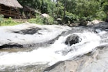 A tranquil riverside scene features a thatched-roof hut nestled among lush green trees and rocks. A gentle stream flows over smooth rocks, creating a small waterfall. The setting is natural and serene, inviting a sense of peace and relaxation.