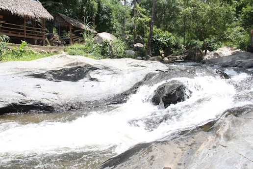A tranquil riverside scene features a thatched-roof hut nestled among lush green trees and rocks. A gentle stream flows over smooth rocks, creating a small waterfall. The setting is natural and serene, inviting a sense of peace and relaxation.