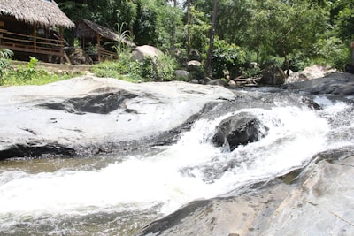 A tranquil riverside scene features a thatched-roof hut nestled among lush green trees and rocks. A gentle stream flows over smooth rocks, creating a small waterfall. The setting is natural and serene, inviting a sense of peace and relaxation.