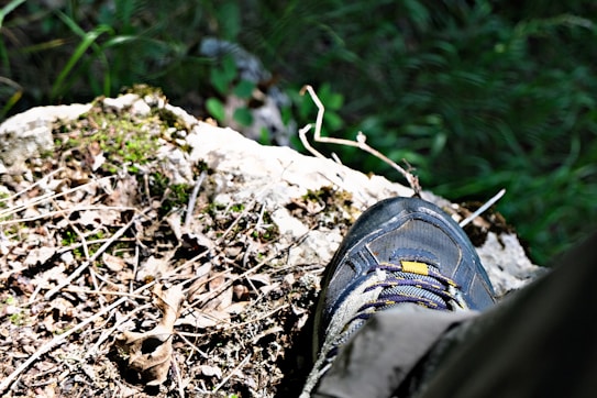A hiking shoe is stepping on a rocky, uneven surface covered with dried leaves, twigs, and patches of green moss. Surrounding the shoe are various shades of green, indicating dense foliage or a forest environment.