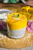 A glass filled with a layered dessert, featuring a chia seed pudding base topped with bright mango puree, garnished with fresh mint leaves and sprinkled coconut. The glass is placed on a wooden board, surrounded by scattered coconut flakes, a few pink flowers, and pieces of diced mango in the background.