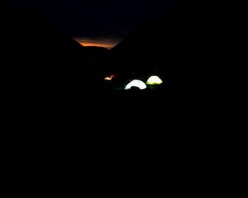 Two glowing tents are set up in a dark outdoor landscape, possibly a campsite at dusk or night. The sky shows a faint orange glow of twilight or sunset between two dark silhouettes of mountains.