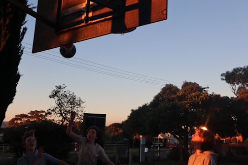 Children playing basketball in an outdoor court during sunset. A basketball is in mid-air near the hoop, and two children are looking up at it while a third prepares to catch or shoot. The backdrop includes trees silhouetted against the evening sky, adding a serene ambiance to the scene.