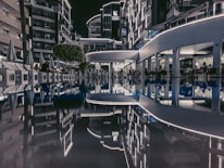 The image depicts an outdoor area of a modern hotel complex at night, with neon lights illuminating the buildings. A pool reflects the surroundings, creating a symmetrical and serene view. People are seated around the area, likely at a restaurant or bar.