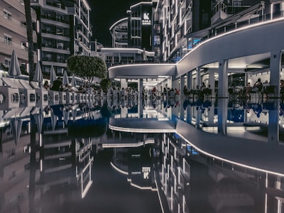 The image depicts an outdoor area of a modern hotel complex at night, with neon lights illuminating the buildings. A pool reflects the surroundings, creating a symmetrical and serene view. People are seated around the area, likely at a restaurant or bar.