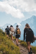 A group of people hiking on a grassy mountain trail with backpacks. They are walking uphill toward snow-capped peaks under a partly cloudy sky. The landscape is lush with green grass and wildflowers.