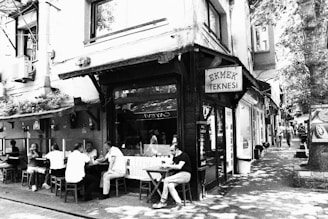 A quaint street scene depicts several people seated outside a small café under a sign reading 'Ekmek Teknesi'. The surrounding area includes a mix of buildings, some with foliage on them, and dappled sunlight filtering through street-side trees.