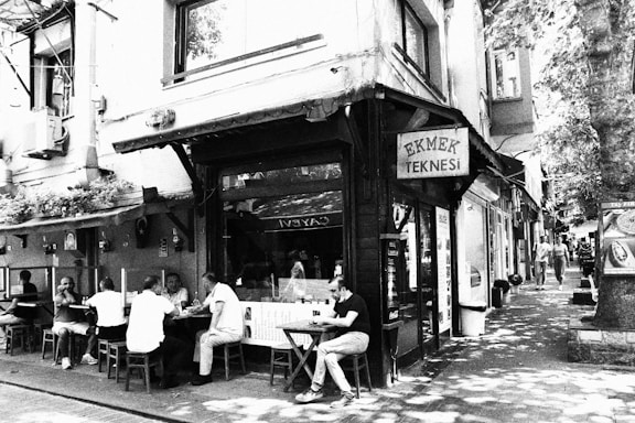 A quaint street scene depicts several people seated outside a small café under a sign reading 'Ekmek Teknesi'. The surrounding area includes a mix of buildings, some with foliage on them, and dappled sunlight filtering through street-side trees.