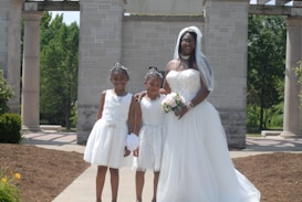 A woman in a bridal gown stands with two young girls wearing white dresses and tiaras. The woman holds a bouquet and wears a veil, while the girls appear to be flower girls. They are posed outside in front of a stone structure with columns and green trees in the background.