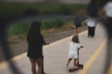 A young child wearing a white outfit is riding a red scooter on a paved path that is bordered by yellow lines. An older child with long dark hair is also riding a scooter nearby. The background shows more people walking along the pathway, with greenery lining the edges.
