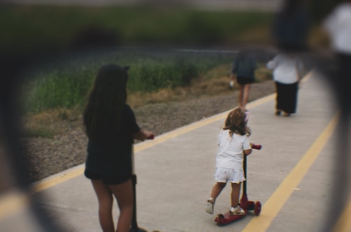 A young child wearing a white outfit is riding a red scooter on a paved path that is bordered by yellow lines. An older child with long dark hair is also riding a scooter nearby. The background shows more people walking along the pathway, with greenery lining the edges.