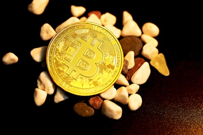 A physical representation of a Bitcoin sits among scattered small, smooth stones on a dark surface. The Bitcoin is prominently placed in the center, showcasing its intricate design and golden color.