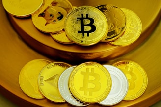 A collection of physical coins representing various cryptocurrencies is arranged on a golden surface. The coins prominently display logos like Bitcoin and Dogecoin, with a mix of gold and silver colors.