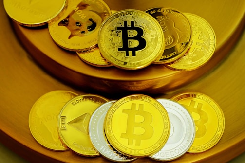 A collection of physical coins representing various cryptocurrencies is arranged on a golden surface. The coins prominently display logos like Bitcoin and Dogecoin, with a mix of gold and silver colors.
