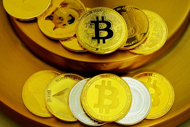 A collection of physical coins representing various cryptocurrencies is arranged on a golden surface. The coins prominently display logos like Bitcoin and Dogecoin, with a mix of gold and silver colors.