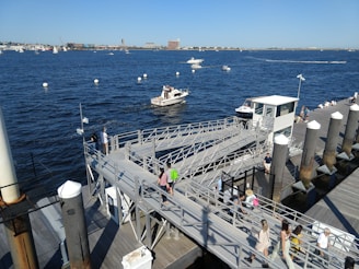A bustling marina with several people walking along a pier and ramps. A boat is cruising on the deep blue water, dotted with buoys and small white boats. Buildings can be seen in the background under a clear blue sky.