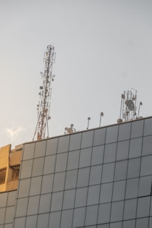 A modern building topped with various antennas and satellite dishes, including a tall metal communication tower. The building features a grid-like facade of glass panels, reflecting the soft light of a setting or rising sun.