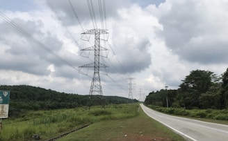 A rural landscape featuring a long, straight road flanked by lush green vegetation on both sides. Tall electricity pylons carrying numerous power lines stretch into the distance, under a sky filled with large, fluffy clouds.