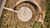 An aerial view of a circular helipad located in an open, grassy area. The helipad is marked with an 'H' in the center and surrounded by a circular path that has worn a darker ring in the grass. A straight path leads from the lower left toward the helipad. Shadows and sunlight create interesting textures on the ground.
