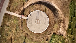 An aerial view of a circular helipad located in an open, grassy area. The helipad is marked with an 'H' in the center and surrounded by a circular path that has worn a darker ring in the grass. A straight path leads from the lower left toward the helipad. Shadows and sunlight create interesting textures on the ground.