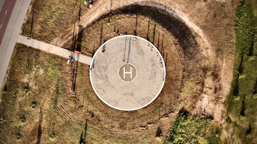An aerial view of a circular helipad located in an open, grassy area. The helipad is marked with an 'H' in the center and surrounded by a circular path that has worn a darker ring in the grass. A straight path leads from the lower left toward the helipad. Shadows and sunlight create interesting textures on the ground.